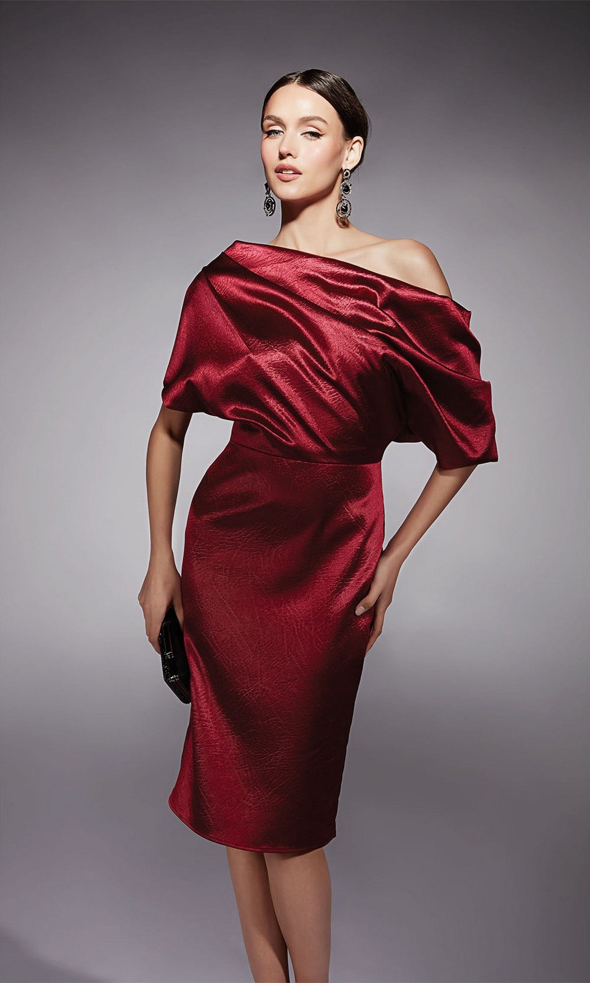 A woman wearing the Knee Length Semi-Formal Dress: Alyce 70060, an off-the-shoulder, shiny cocktail dress with short sleeves, styled with a black clutch and dangling earrings.